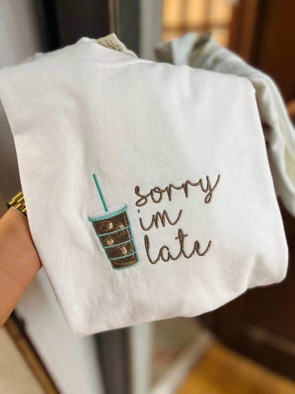 A white fabric with embroidered text 'Sorry I'm late' and a stylized iced coffee design held in a hand.